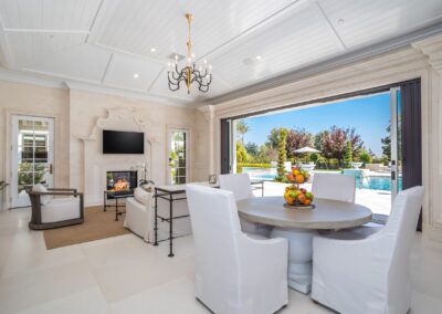 A luxurious living room with white furniture, a round dining table, and a fireplace. Glass doors open to a patio with a pool and garden view.