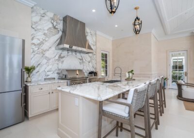 Modern kitchen with marble countertops and backsplash, stainless steel appliances, and a center island with high chairs. Two pendant lights hang from the ceiling. A door leads to an outdoor area.