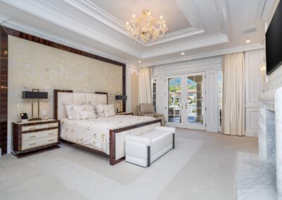 Luxurious bedroom with a large bed, ornate chandelier, and French doors opening to a balcony. Neutral tones with decorative wallpaper and elegant furnishings.