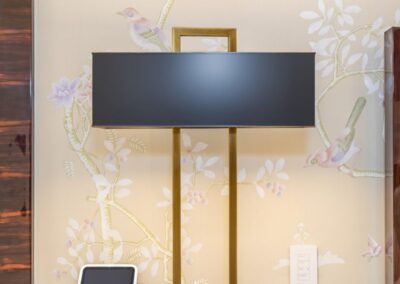 A modern, rectangular black and gold table lamp on a wooden surface next to a tablet and a small gold clock, with floral wallpaper in the background.