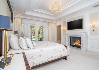 Luxurious bedroom with a large bed, elegant chandelier, fireplace, and a flat-screen TV. French doors open to an outdoor view. Cream and white decor with floral bedding.