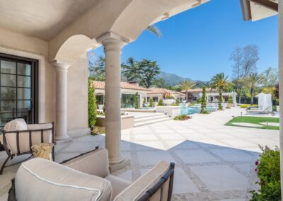 Luxury patio with plush seating overlooks a spacious courtyard featuring a pool, greenery, and mountain views under a clear blue sky.