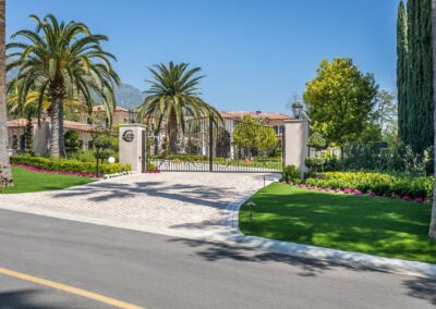 Gated entrance of a luxurious property with palm trees, manicured lawns, and vibrant flowerbeds under a clear blue sky.