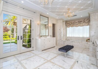 Luxurious marble bathroom with a large window, chandelier, a freestanding tub, and a black bench. White double doors open to a garden view.