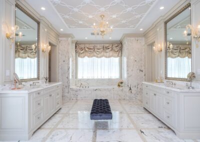 Luxurious marble bathroom with dual sinks, a central navy bench, elegant chandeliers, and large mirrors. Draped window above a spacious bathtub.