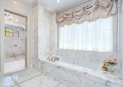 Luxurious marble bathroom with a large tub, elegant drapery above the window, and a walk-in shower visible through an open door.