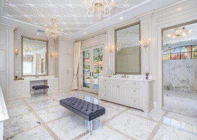 Luxurious white marble bathroom with two large mirrors, chandeliers, and a central tufted bench. Double doors lead to an outdoor area, and a glass-enclosed shower is visible on the right.