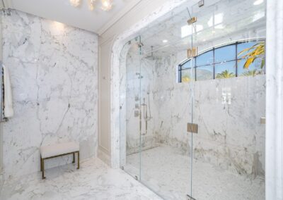 Luxurious marble bathroom with a large glass-enclosed shower, a bench, and a window view of palm trees.