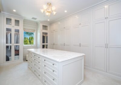 A spacious walk-in closet with white cabinetry, a large marble-topped island, and a hanging chandelier. Light streams in from a window with open blinds.