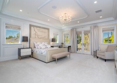 Elegant bedroom with a large bed, floral bedding, two nightstands, a chandelier, and French doors leading to a balcony. The room is well-lit and features a neutral color palette.