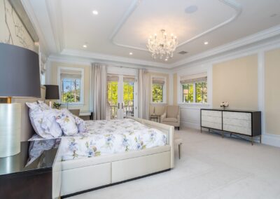 Elegant bedroom with a chandelier, floral bedding, two windows with curtains, an armchair, and a dresser. Walls are a soft beige with white trim.