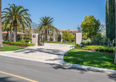 A gated entrance to a luxurious estate with palm trees, manicured lawns, and colorful flowerbeds under a clear blue sky.