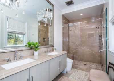 Modern bathroom with a double sink vanity, large mirror, glass-enclosed shower, marble flooring, chandelier, and a window with a Roman shade.