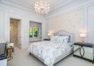 Elegant bedroom with a plush bed, floral bedding, chandelier, and two mirrored nightstands. Soft lighting and a window with a view create a bright and inviting atmosphere.