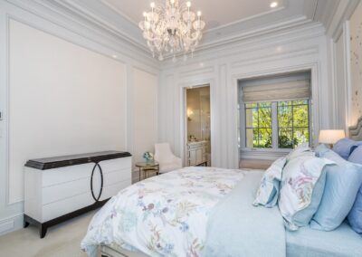 Elegant bedroom with a floral-patterned bed, white dresser, and chandelier. A large window offers a view of greenery, and an open door leads to a bathroom.