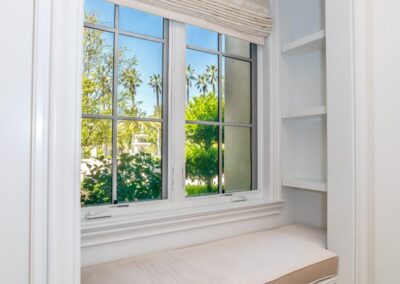 A bright window seat with a beige cushion and white drawers beneath, surrounded by shelves, overlooks greenery and palm trees outside.