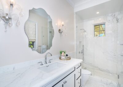 A modern bathroom with a marble countertop, sink, ornate mirror, and walk-in shower. The décor includes wall sconces and a small decorative plant basket.