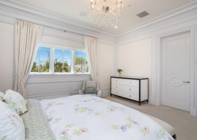 A bright bedroom with a floral bedspread, a white dresser, and a light gray armchair. Large windows with curtains let in natural light. A chandelier hangs from the ceiling.