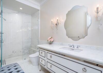 Elegant bathroom with a marble sink, ornate mirror, and glass-enclosed shower. Soft lighting from wall sconces, and a small bouquet on the counter adds a touch of color.