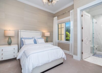 Elegant bedroom with a white bed, two bedside tables with lamps, large window, and an open door leading to a marble-tiled bathroom.