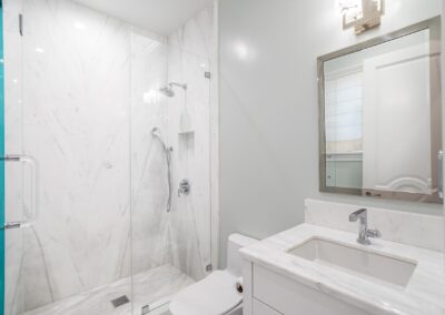 A modern bathroom with a glass-enclosed shower, a white sink with a chrome faucet, and a wall-mounted mirror. Walls are a light gray color, and the shower has white marble tiles.