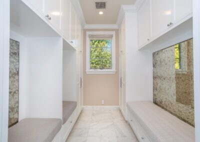 A modern room with white walls and cabinets, cushioned benches, and a large window. The floor is tiled in a marble pattern.