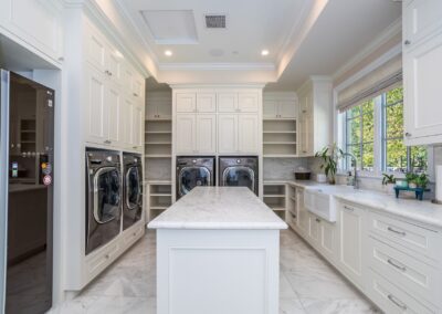 A spacious white kitchen with marble countertops, built-in ovens, a central island, and large windows overlooking greenery.