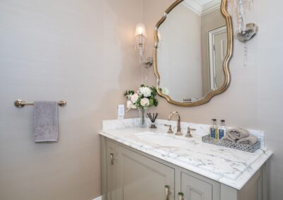 Bathroom with a marble countertop, ornate mirror, wall sconce, and floral arrangement. Towels are placed on the counter next to soap dispensers. Beige walls and a hanging hand towel complete the decor.