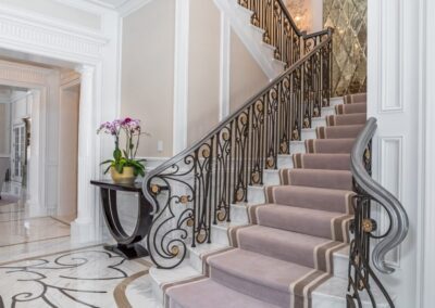 A grand staircase with ornate iron railings, lavender carpet, and a decorative marble floor. There is a table with potted orchids at the base.