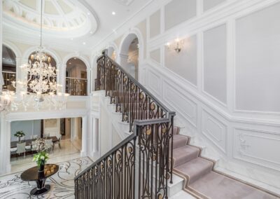 A grand, opulent staircase with ornate railing, leading to an upper level. Features a large chandelier, white walls, and marble flooring with intricate patterns.