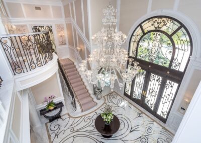 Opulent foyer with a grand staircase, ornate chandelier, and elegant decor featuring a round table and floral arrangement. Large arched window with intricate detailing above the entrance.