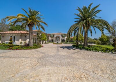 A large, luxurious house with a paved circular driveway is surrounded by palm trees and landscaped gardens under a clear blue sky.