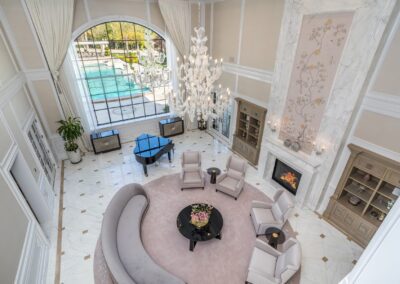 Luxurious living room with high ceiling, chandelier, grand piano, curved sofa, fireplace, and large arched window overlooking a swimming pool.