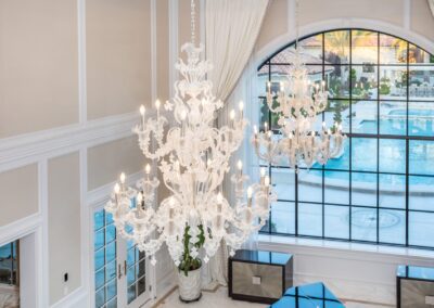 Luxurious room with two ornate white chandeliers, large arched window, and view of a pool outside.