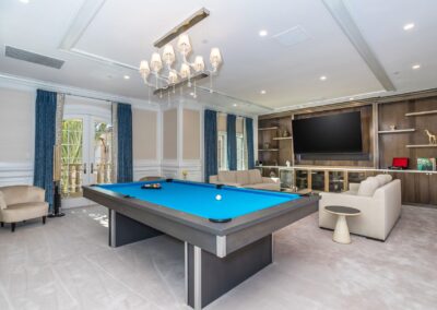 A spacious game room featuring a blue pool table, beige couches, a large TV, and shelves with decorative items. Blue curtains adorn the windows.