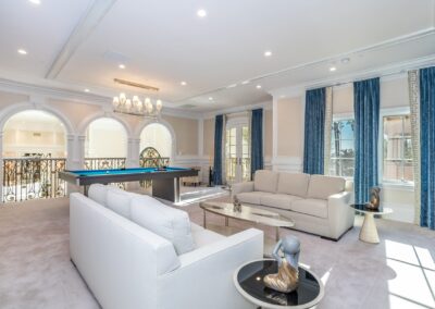 Spacious living room with white sofas, glass tables, a pool table, large windows with blue curtains, white walls, and a chandelier.