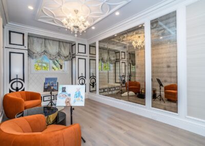 Elegant room with mirrored walls, orange chairs, a small round table, and a chandelier. An easel with a painting and a window with ornate drapes are also present.