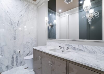 Luxurious bathroom with a marble wall and countertop, a large mirror, modern sink fixtures, and a white toilet. Wall-mounted light fixtures are on either side of the mirror.