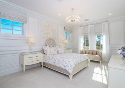 A bright, elegant bedroom with a floral-patterned bed, a tufted bench, two side tables with lamps, a chandelier, and a vase with flowers on the dresser. White walls and large windows with curtains.