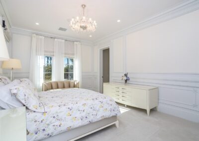 A bright bedroom with a floral-patterned bed, chandelier, dresser, and a small sofa. The room has large windows with curtains, white walls, and decorative molding.