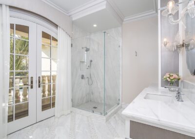 Elegant bathroom with marble flooring, a glass shower, and a white vanity. French doors with curtains open to a balcony with palm trees in view. Wall-mounted lights illuminate the space.