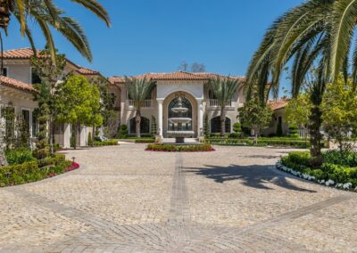 Large mansion with red-tiled roof and central archway, surrounded by palm trees and a cobblestone driveway with a fountain in front.