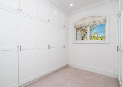 A bright, empty walk-in closet with white cabinetry and a small window with a beige shade, overlooking greenery outside.