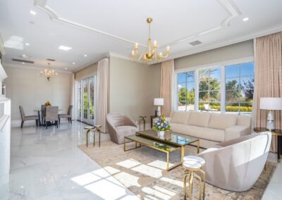 A spacious, elegant living room with beige sofas, a glass coffee table, chandeliers, large windows, and a view of a dining area.