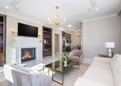 Elegant living room with a lit fireplace, wall-mounted TV, beige sofa, glass coffee table with flowers, and modern chandelier. Kitchen area visible in the background.