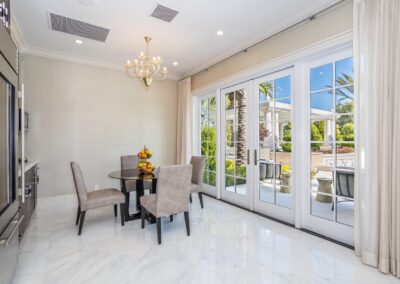 A dining area with a round table and four chairs on a marble floor, adjacent to large glass doors opening to a patio with outdoor seating. A chandelier hangs from the ceiling.