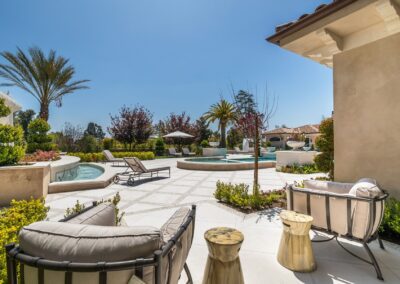 Luxurious outdoor patio with cushioned seating, small round tables, pool, and palm trees under a clear blue sky.