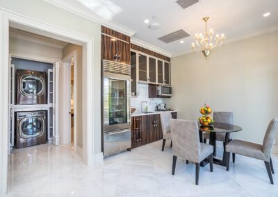 Modern kitchen with brown cabinets, stainless steel fridge, glass dining table with four chairs, and a chandelier. Laundry area visible through an open door on the left.