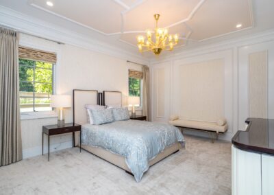 A bedroom with a double bed, padded headboard, two nightstands with lamps, a bench, and a chandelier. The room has light-colored walls, carpet, and two windows with curtains.