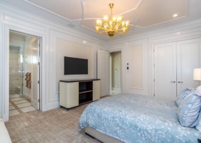 A modern bedroom with a bed featuring blue floral bedding, a wall-mounted TV, a chandelier, and an open door leading to a bathroom. White walls and carpet complete the room.